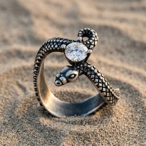 May include: A silver-toned metal ring shaped like a coiled snake with a textured, scale-like pattern along the band. The snake's head is positioned near a single, round, clear faceted gemstone held in a prong setting at the top of the ring.