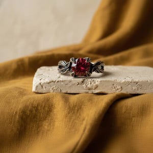 May include: A ring featuring a round, faceted deep red gemstone set in a dark, oxidised metal band. The band is crafted with intricate, leaf-like filigree detailing that extends from the central setting, creating a textured and ornate appearance.