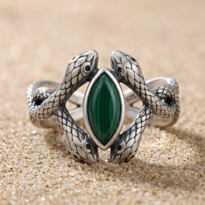 Könnte beinhalten: Ein Ring aus Sterlingsilber mit zwei detaillierten Schlangen, deren texturierte Schuppen in der Mitte aufeinandertreffen. Zwischen den Schlangenköpfen befindet sich ein marquise-geschliffener grüner Malachit. Die Zahl 925 ist innen eingeprägt.