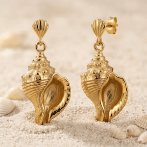 May include: A pair of gold-toned drop earrings featuring a detailed conch shell design. Each earring has a small, fan-shaped post top connected to a textured, spiraled seashell pendant that hangs vertically from a circular jump ring.