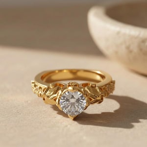 May include: A gold ring with a large, clear, round-cut diamond center stone. The ring has intricate floral and leaf detailing on the band and around the diamond. The ring is set on a neutral surface.