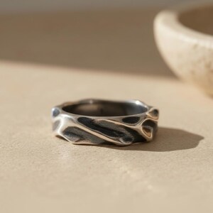 Puede incluir: Un anillo de plata con un diseño texturizado en forma de ola. El anillo tiene un acabado oscuro oxidado, creando contraste con las zonas más claras y pulidas. La banda es gruesa y sustancial, con un patrón orgánico y fluido. Joyería.