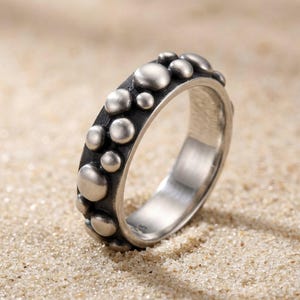 Könnte beinhalten: Ein Ring aus silberfarbenem Metall mit einem dunklen, oxidierten Band, das auf der Außenseite mit zahlreichen erhabenen, glatten, kreisförmigen Kugeln unterschiedlicher Größe verziert ist, was eine texturierte, blasenartige Optik mit polierter Innenseite erzeugt.