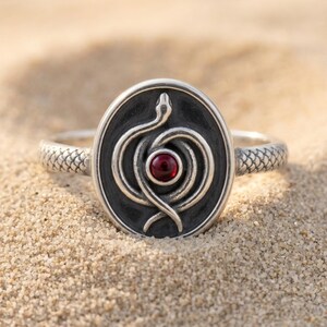 Peut inclure: Une bague en argent avec un plateau ovale orné d'un serpent en relief enroulé autour d'une pierre précieuse rouge ronde. L'anneau présente un motif d'écailles texturé et le fond du plateau ovale est oxydé pour une finition gris foncé.