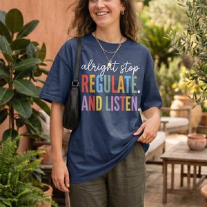 May include: A navy blue t-shirt with the text "alright stop REGULATE. AND LISTEN." in colorful block letters. The shirt is worn with olive green pants. The words "True Navy" are in the upper left corner.