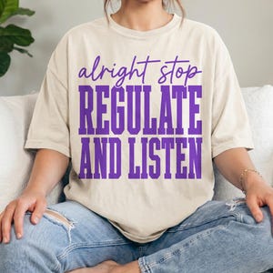 Può includere: T-shirt color avorio con la scritta "alright stop REGULATE AND LISTEN" in lettere viola. La maglietta è indossata con jeans blu. Il tema generale è l'abbigliamento casual.