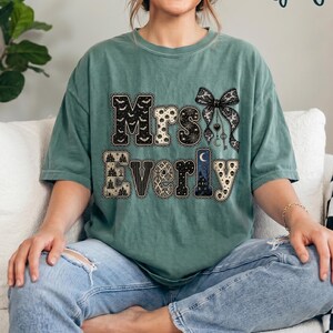 May include: A light green short-sleeve t-shirt featuring the text "Mrs Everly" in a decorative font. The letters are filled with various patterns including bats, stars, skulls, and a haunted house scene. A black lace bow graphic is above the text.