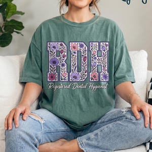 May include: A light green short-sleeve t-shirt featuring the letters RDH in a large block font filled with purple, pink, and white floral illustrations. Below the letters, the text Registered Dental Hygienist is printed in a white script font.
