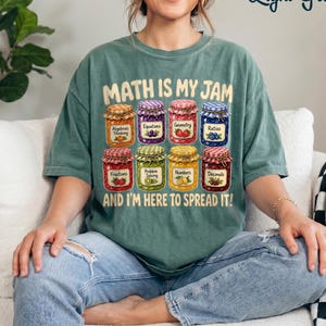 May include: A light green t-shirt featuring a graphic of eight colorful jars of jam, each labeled with a math topic. The text reads "MATH IS MY JAM AND I'M HERE TO SPREAD IT!" The jars contain various fruits and are arranged in two rows of four.