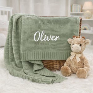 以下が含まれることがあります： 白で「Oliver」と刺繍されたセージグリーンのニットベビーブランケット。柳のバスケットに入れられ、クリーム色のリボンが付いたぬいぐるみキリンが隣にあり、居心地の良い保育園のテーマを演出しています。