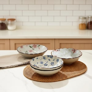 Könnte beinhalten: Drei Keramikschalen mit floralen Mustern. Eine Schale hat rosa Blüten, eine andere blaue Blüten und die dritte ein subtiles Blumenmuster. Die Schalen stehen auf einem gewebten Platzset, was auf ein Ess- oder Küchenthema hindeutet.