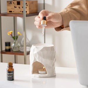 以下が含まれることがあります： テクスチャ加工された表面とティーライトキャンドルの小さな開口部を備えた、白いセラミックエッセンシャルオイルディフューザー。人がスポイトを使ってオイルを加えています。フレグランスオイルの小さな琥珀色のボトルがディフューザーの隣にあります。