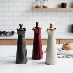 May include: Three ceramic oil and vinegar dispensers in a kitchen setting. The dispensers are in black, burgundy, and off-white. Each has a narrow neck, a spout, and a small handle on top. The background includes a white tile backsplash and a loaf of bread.