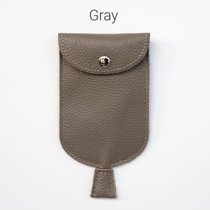 Op de afbeelding: Een grijs, getextureerd tasje met een afgeronde flap en een zilverkleurige metalen drukknoopsluiting. Het tasje heeft een klein, rechthoekig lipje dat uit het midden aan de onderkant steekt. Het woord Gray staat in zwarte tekst erboven.