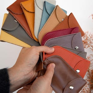 Puede incluir: Una colección de pequeñas fundas de cuero dispuestas en abanico, con varios colores que incluyen marrón, naranja, rojo, morado, azul, amarillo y beige. Cada funda tiene una solapa redondeada asegurada con un botón de presión de metal plateado.