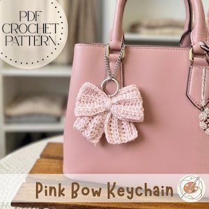 Könnte beinhalten: Ein hellrosa gehäkelter Schleifen-Schlüsselanhänger, der an einer rosa Handtasche hängt. Die Schleife ist an einer silbernen Kette und einem Ring befestigt. Der Text "Pink Bow Keychain" steht unten, mit "PDF CROCHET PATTERN" oben.