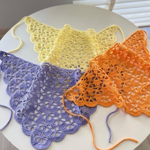 May include: Four handmade crochet neck scarves in yellow, purple, and orange. Each scarf has a scalloped edge and a tie closure. The openwork design is visible in the close-up shot. These accessories are perfect for adding a touch of handmade charm to any outfit.