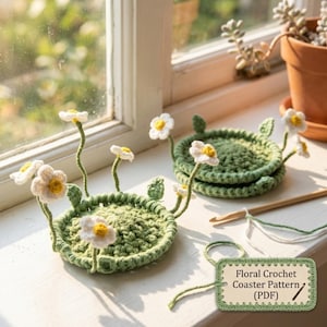 Puede incluir: Dos posavasos de ganchillo de color verde salvia con flores blancas de centro amarillo sobre tallos finos. Una etiqueta rectangular con borde verde muestra el texto: Floral Crochet Coaster Pattern (PDF). Un ganchillo de madera está cerca.