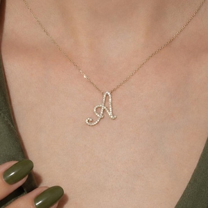 Puede incluir: Un collar de cadena dorada con un colgante en forma de letra A cursiva. El colgante está adornado con pequeñas piedras claras y brillantes incrustadas a lo largo de toda la superficie de la letra, creando un aspecto delicado y refinado sobre la piel.