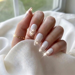 May include: A hand with almond-shaped nails featuring a white base colour and silver glitter tips. The nails have a glossy finish and a sparkling effect. A thin gold chain-link ring is worn on the ring finger, adding a metallic accent to the look.
