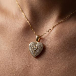 Close-up of a dainty pavé heart pendant necklace on a delicate gold chain. The 925 sterling silver heart is covered in small sparkling stones with a tiny raised heart detail and fine center line, shown on a woman’s neck and in hand, minimalist jewelry style, ideal Valentines gift for her.