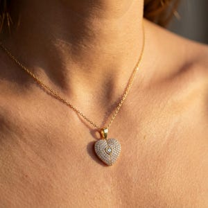 Puede incluir: Un collar dorado con un colgante en forma de coraz&oacute;n. El colgante est&aacute; incrustado con peque&ntilde;as piedras transparentes y presenta una piedra en forma de coraz&oacute;n m&aacute;s peque&ntilde;a en el centro. El collar se lleva alrededor del cuello.