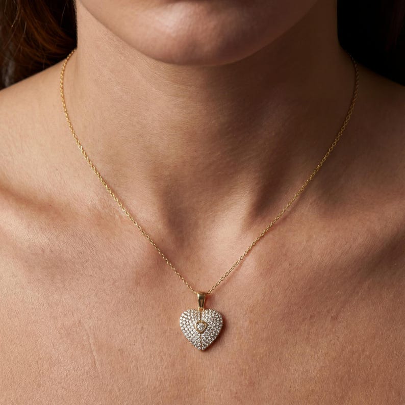 Puede incluir: Un collar dorado con un colgante en forma de coraz&oacute;n. El colgante est&aacute; adornado con peque&ntilde;as piedras preciosas transparentes, con un coraz&oacute;n m&aacute;s peque&ntilde;o en el centro. El collar se muestra en el cuello de una persona.