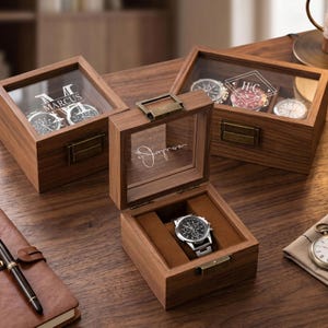 May include: Three wooden watch boxes with glass lids and brass-toned latches. One box is open, displaying a silver chronograph watch on a brown cushion. The lids feature personalized engravings including the names Marcus, Julian, and the initials H C.
