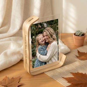 May include: A wooden photo frame with a clear acrylic panel displays a photo of a woman and a child. The frame has a curved design and the text "Forever my Mom, forever my friend" is engraved on the side. The frame sits on a wooden surface.