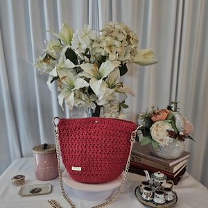 May include: A red crochet bucket bag with a gold chain strap. The bag has a small rectangular tag that says "handmade". The bag is sitting on a white surface, with a bouquet of white flowers in the background.