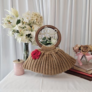 May include: A tan crocheted garment with a red rose accent, displayed on a white surface. The garment is surrounded by decorative elements, including a vase of white flowers, a pink jar, and a small vase of dried roses.