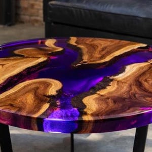 May include: A round coffee table with a unique design. The tabletop features a vibrant purple resin river flowing between natural wood sections. The table is supported by black metal legs, creating a modern aesthetic.