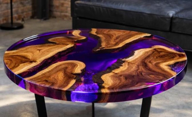 May include: A round coffee table with a unique design. The tabletop features a vibrant purple resin river flowing between natural wood sections. The table is supported by black metal legs, creating a modern aesthetic.