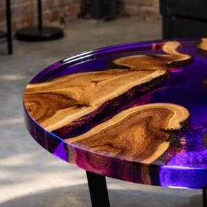 May include: A round wooden table with a dark purple epoxy resin top. The table features natural wood grain patterns and a glossy finish. The table has black metal legs and is suitable for indoor use.