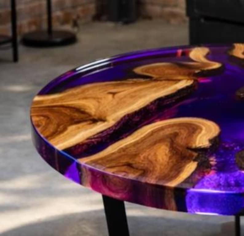 May include: A round wooden table with a dark purple epoxy resin top. The table features natural wood grain patterns and a glossy finish. The table has black metal legs and is suitable for indoor use.