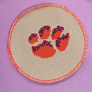 May include: A round, orange and purple embroidered patch with a paw print design. The patch has a white background and a red stitched border.