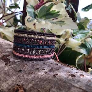 Peut inclure: Un large bracelet tissé dans les tons marron, sarcelle, rose et or. Le bracelet est fait d'une matière tissée et repose sur un morceau de bois. Le bracelet a un design unique avec de multiples couches et textures.