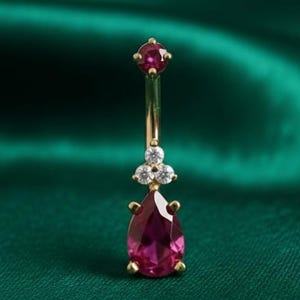 May include: A gold-colored belly button ring with a teardrop-shaped, deep pink gemstone at the bottom. The ring features a small, round pink gemstone at the top and three small, clear gemstones in the middle. The background is a dark green fabric.