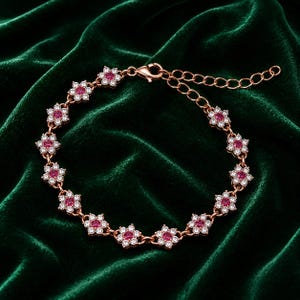 May include: A rose gold tone bracelet featuring a series of connected flower-shaped links. Each flower link contains a central round red stone surrounded by six small clear white stones. The bracelet includes a lobster claw clasp and an extender chain.