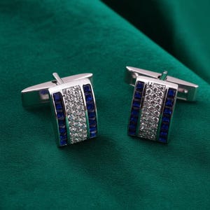 May include: A pair of rectangular silver-toned cufflinks featuring a central vertical row of clear, sparkling stones flanked by two vertical rows of deep blue square-cut stones. The metal finish is polished and reflective, with a standard hinged toggle back.