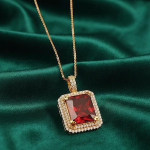 May include: A gold necklace featuring a rectangular pendant with a large, deep red center stone. The pendant is framed by two rows of small, clear, sparkling stones. The gold chain is thin and delicate, connecting to the top of the pendant.