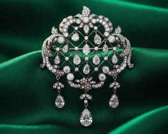 May include: A dark metal brooch featuring an intricate lattice design adorned with numerous clear, round-cut stones and several dangling, pear-shaped clear stones. The piece has a symmetrical, ornate structure with a polished, dark finish.