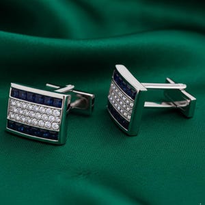 Peut inclure: Une paire de boutons de manchette rectangulaires argentés avec une bande centrale horizontale de pierres claires rondes, encadrée par deux rangées horizontales de pierres carrées bleu foncé. La monture métallique est polie avec un fermoir basculant.