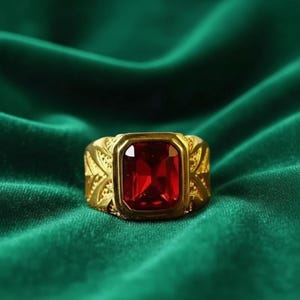 May include: A gold-toned ring featuring a large, rectangular, faceted red gemstone set in the center. The band of the ring displays an intricate, textured leaf-like pattern on both sides of the central stone setting, creating a detailed design.