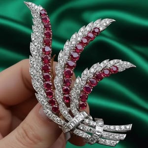 May include: A silver-toned brooch shaped like a spray of leaves, featuring three prominent curved stems set with round, deep red rubies. The leaves are encrusted with small, clear, sparkling stones, and the base is tied with a band of baguette-cut stones.