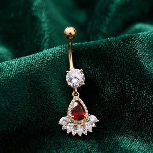 May include: A gold-toned curved barbell belly ring featuring a round clear cubic zirconia stone at the top and a dangling pear-shaped red stone pendant surrounded by a halo of smaller clear stones and a fan-like arrangement of marquise-cut stones.