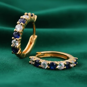 May include: A pair of gold hoop earrings featuring an alternating pattern of round-cut blue sapphire-colored stones and clear white stones. The hoops have a hinged closure mechanism and are designed with a polished gold metal finish throughout.