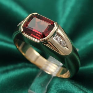 May include: A gold ring with a large, rectangular, red gemstone as the centerpiece. The ring has a gold band and gold accents around the red stone, with small diamonds on the side. The ring is displayed on a clear stand.