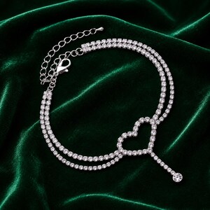 May include: A silver-toned double-strand anklet featuring a central heart-shaped charm encrusted with clear rhinestones. The piece includes a delicate chain extension with a lobster claw clasp and a single dangling rhinestone at the end of the heart.