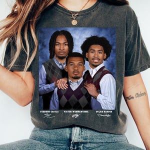 May include: Dark gray t-shirt featuring a printed image of three men in argyle vests and collared shirts. The names Stephon Castle, Victor Wembanyama, and Dylan Harper are printed below the image, along with their signatures.